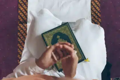 Handen met gebedsparels naast een open heilig boek op houten tafel met ochtendlicht